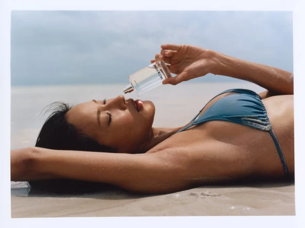 Model Hyunjoo Hwang lies on a sandy beach holding a Salt & Stone fragrance bottle near her face while wearing a teal bikini.