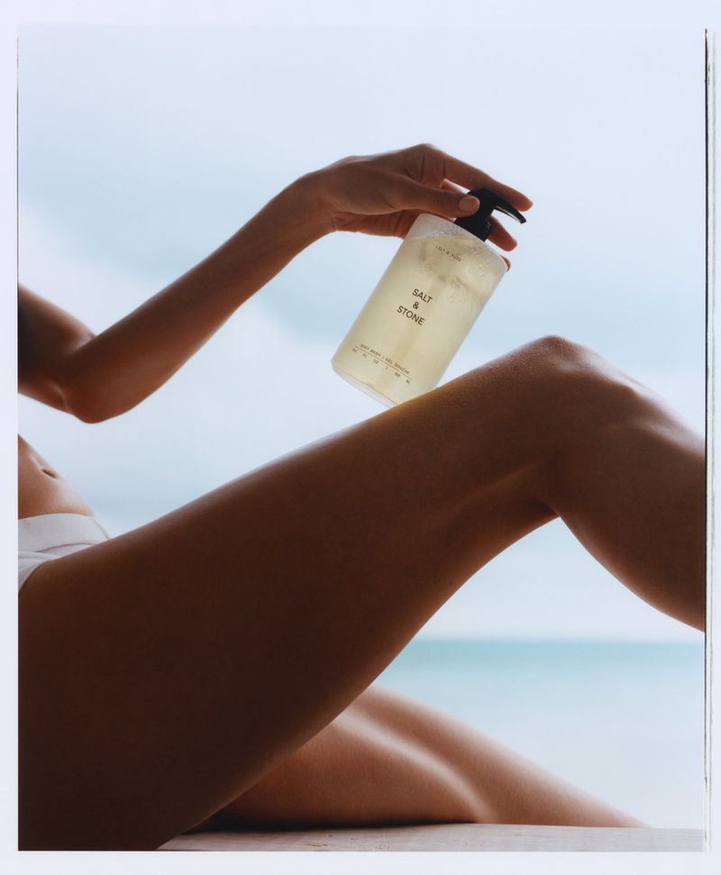 Model Hyunjoo Hwang holding a translucent Salt & Stone pump bottle near her leg against a soft ocean background.