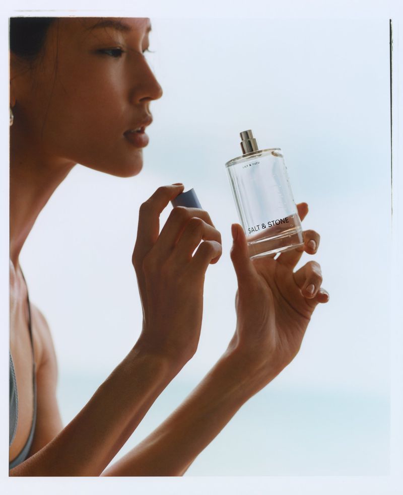 East Asian model Hyunjoo Hwang holds a clear glass Salt & Stone fragrance bottle against a bright open sky.
