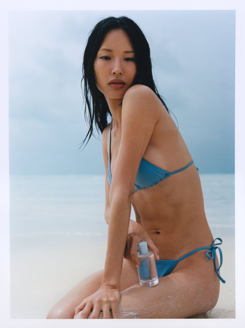 Model Hyunjoo Hwang sitting on a sandy beach wearing a blue bikini and holding a Salt & Stone body mist bottle.