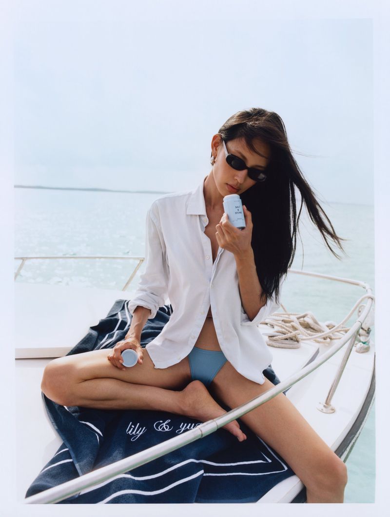 Hyunjoo Hwang wearing a white shirt and sunglasses holding a Salt & Stone deodorant on a boat deck at sea.