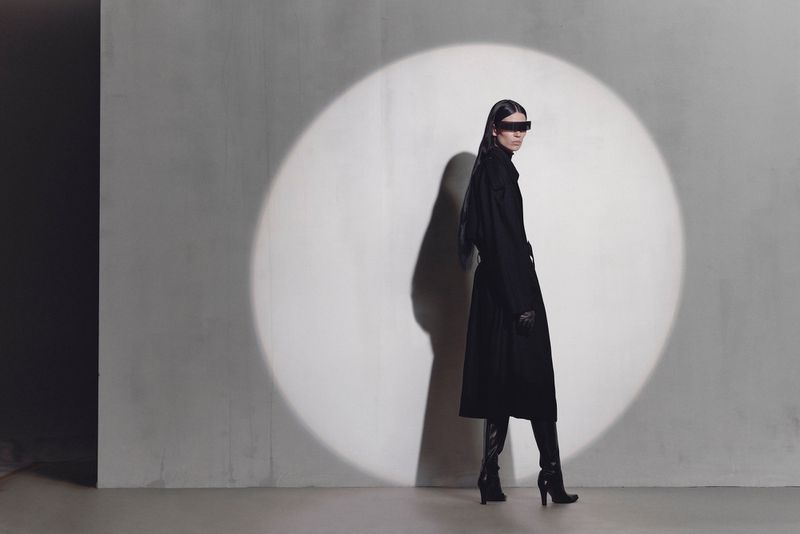 Woman poses in black coat and futuristic visor glasses in a fashion studio, spotlight on a grey textured backdrop, showcasing modern style.