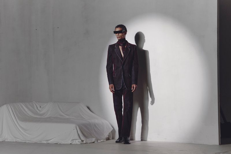 A single adult stands in a photo studio wearing a dark suit and rectangular glasses. He is next to a draped couch and a strong light.