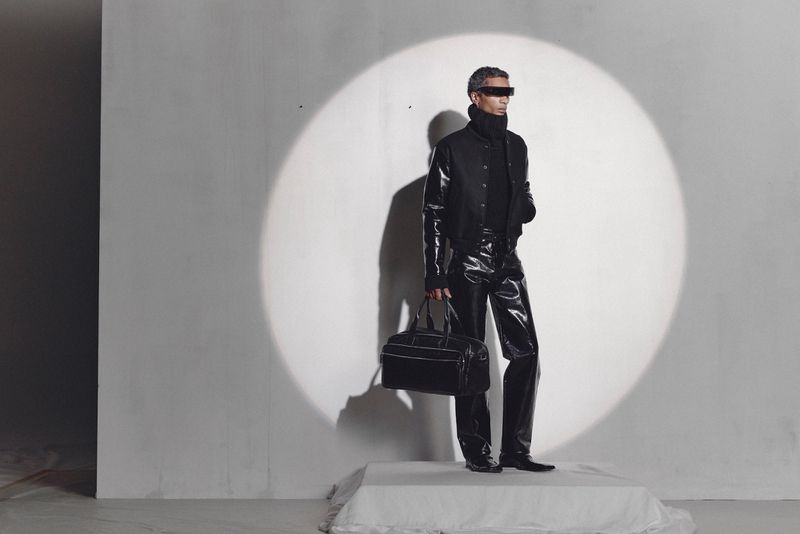 A grey-haired adult man with a modern futuristic fashion style poses with a bag in a light-filled studio wearing dark glasses.