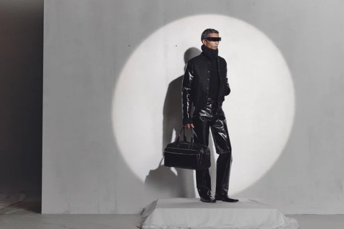 A grey-haired adult man with a modern futuristic fashion style poses with a bag in a light-filled studio wearing dark glasses.