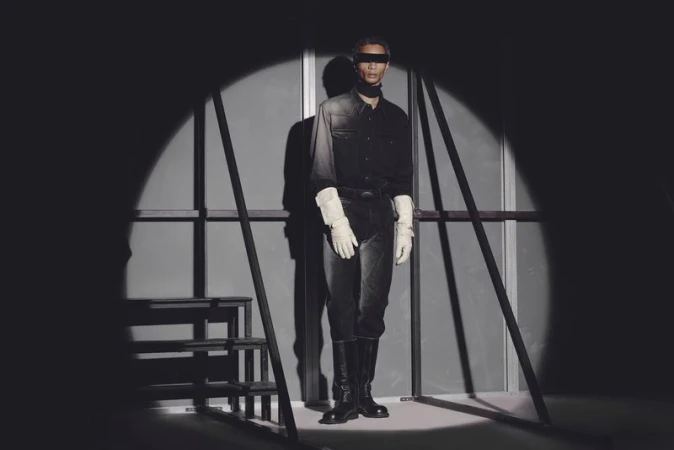 A fashion shot of a model in dark clothing and boots, standing in a spotlight with ladder elements and a monochromatic backdrop.