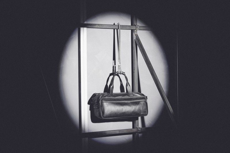 Black leather duffel bag hanging on a metal frame in a spotlit indoor setting, showcasing its design and material under studio lighting.