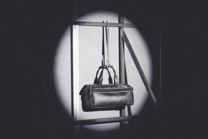 Black leather duffel bag hanging on a metal frame in a spotlit indoor setting, showcasing its design and material under studio lighting.