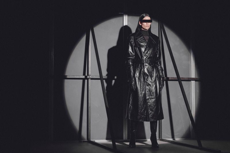 A woman in a black leather trench coat, gloves, and visor poses in front of a gray backdrop, lit by a bright spotlight.