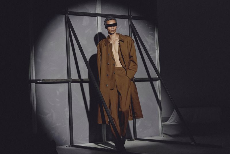Fashion shot of a single adult male model with a brown trench coat and pants stands in a photo studio with spotlight lighting.