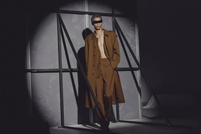 Fashion shot of a single adult male model with a brown trench coat and pants stands in a photo studio with spotlight lighting.