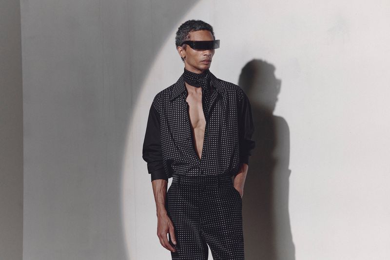 Model wearing dark futuristic visor sunglasses and an open black patterned shirt with matching neck scarf against a neutral wall.