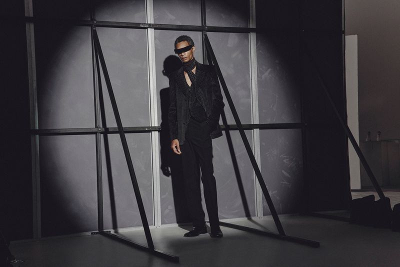 Fashionable man strikes a pose indoors wearing a patterned suit, scarf, and futuristic sunglasses, illuminated by a spotlight.
