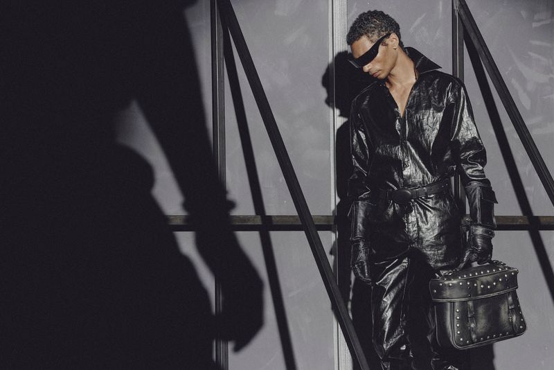 An adult male in a black jumpsuit and sunglasses holds a studded black bag while standing in an indoor setting with a large shadow.