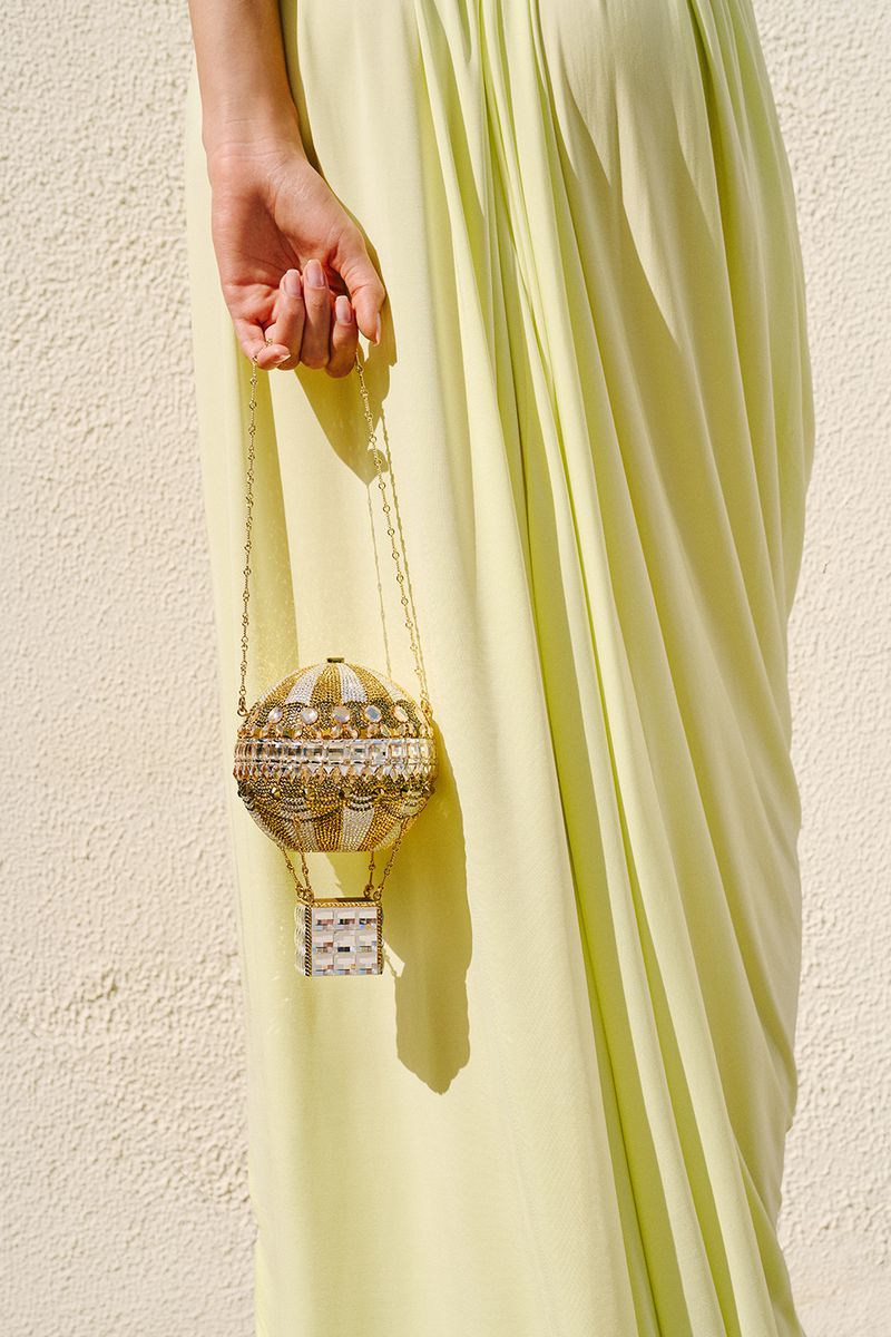 Hand holding an ornate crystal-embellished hot air balloon clutch bag against a pastel yellow draped dress in bright sunlight.