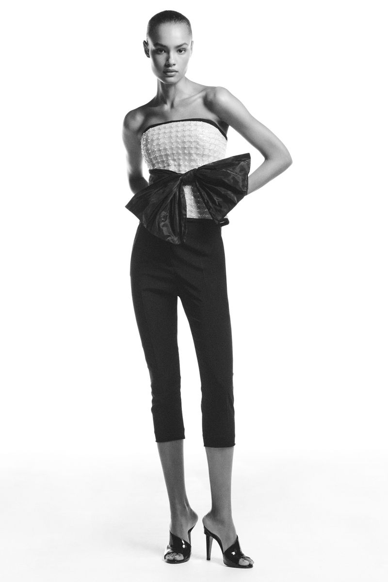 Full-length fashion shot of a slender adult female model wearing a strapless top, black capri pants, and high heels in a white studio.
