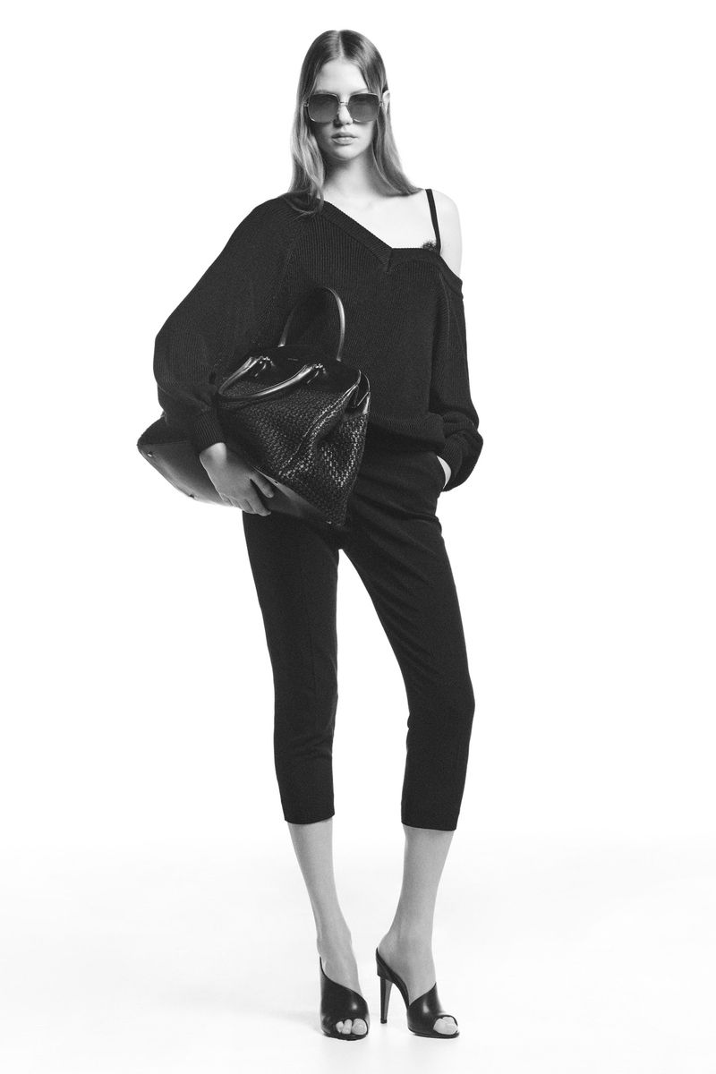 Black-and-white fashion shot of a model in dark clothing holding a woven handbag in a minimalist studio setting with soft lighting.