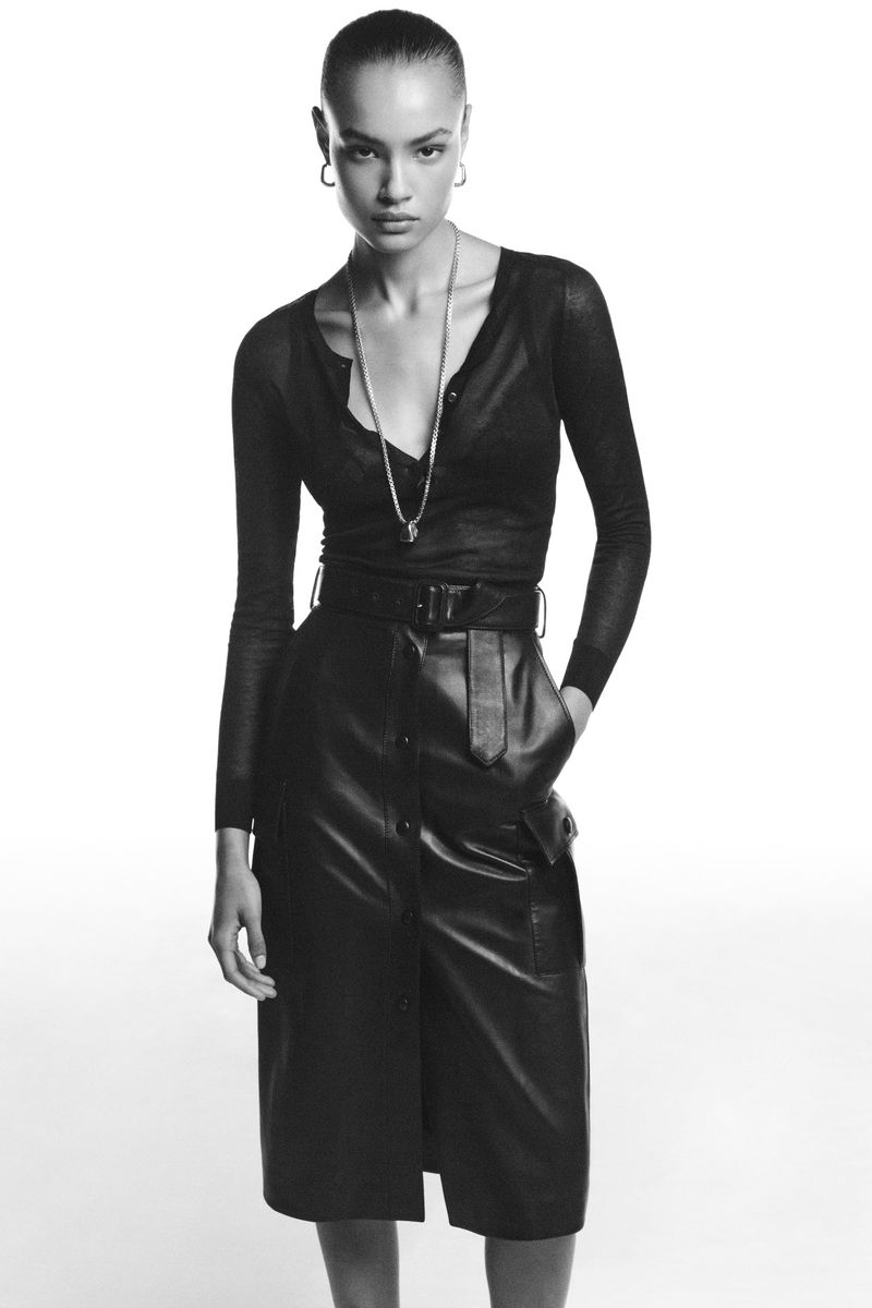 Model wearing a black long-sleeve top and belted black leather midi skirt against a plain white studio background.
