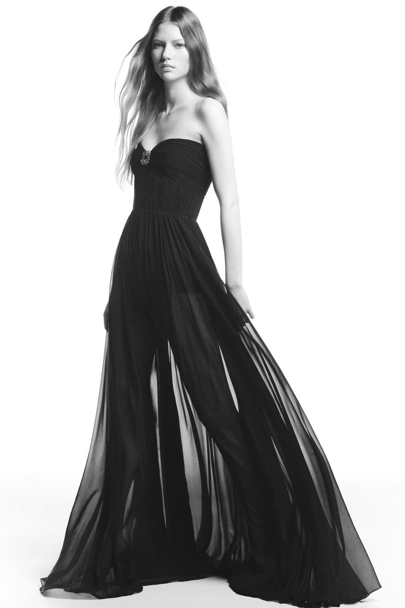 A fair-skinned woman wearing a long black chiffon strapless gown stands in a photo studio, facing forward with a neutral expression.