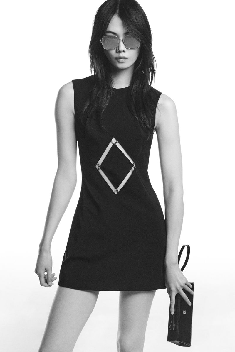 Model wearing a black sleeveless mini dress with a geometric diamond cutout detail and sunglasses against a white studio backdrop.