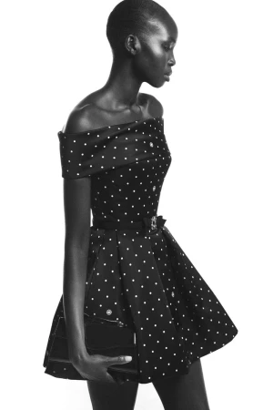 Model Ajok Madel in a black off-the-shoulder polka dot mini dress holding a clutch purse against a white studio background.