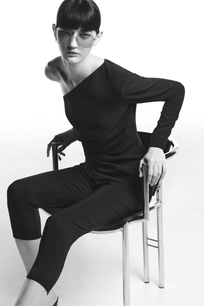 Monochrome fashion shot of a female model in sunglasses and a black jumpsuit, seated on a clear chair against a white background.