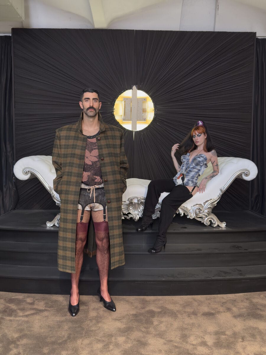 Man in a plaid coat, garter shorts, stockings, and heels standing in front of a woman reclining on an ornate white chaise lounge.