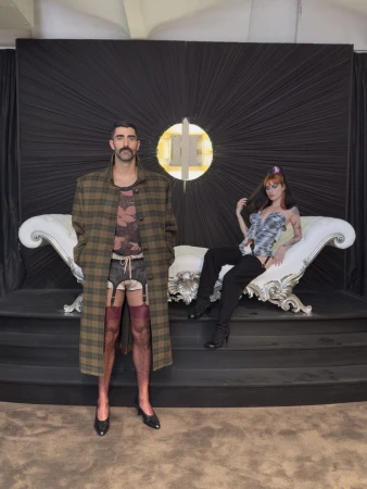 Man in a plaid coat, garter shorts, stockings, and heels standing in front of a woman reclining on an ornate white chaise lounge.