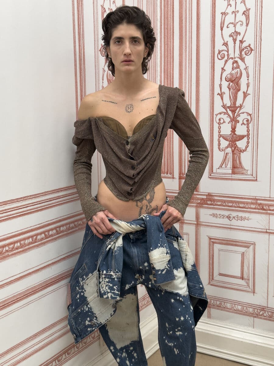 Model wearing a brown off-the-shoulder corset top and bleach-splattered denim jeans against an illustrated wallpaper backdrop.
