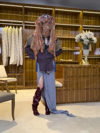 Model wearing a draped mesh head covering, purple shirt, and checkered skirt with tall dark boots inside a walk-in closet