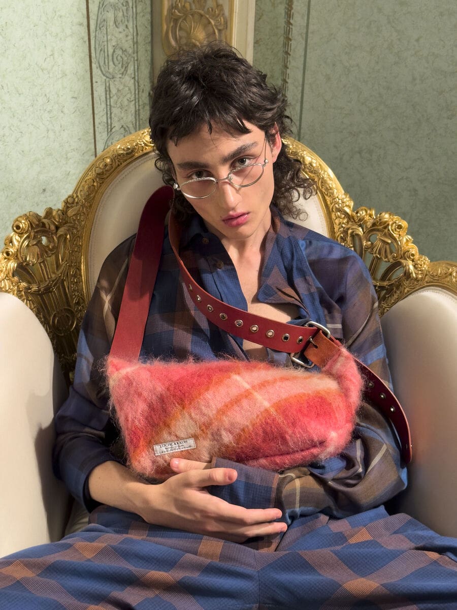 Model in a blue plaid shirt and wire-rim glasses holding a fuzzy pink crossbody bag while seated in a gilded armchair.