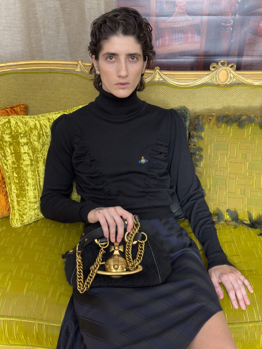 Model in a black turtleneck sweater and striped skirt seated on a yellow velvet couch holding an orb logo handbag.