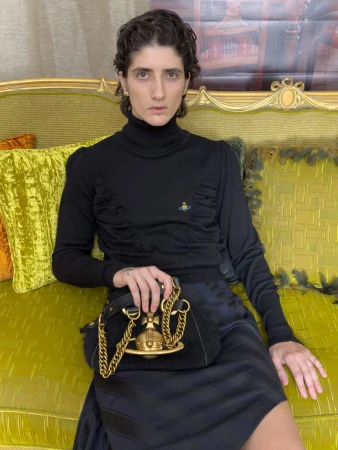 Model in a black turtleneck sweater and striped skirt seated on a yellow velvet couch holding an orb logo handbag.