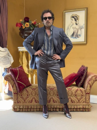 Man wearing a grey suit jacket, satin trousers, patterned shirt, and heeled shoes standing before a sofa