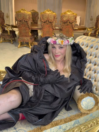 Cicciolina reclines in a voluminous dark hooded cape and pastel flower crown on an ornate gilded tufted chaise longue.