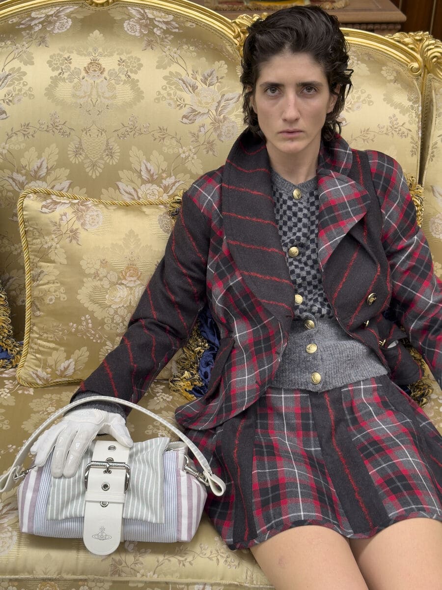 Model in a red and grey plaid suit seated on an ornate gold damask sofa resting a gloved hand on a striped Vivienne Westwood handbag.