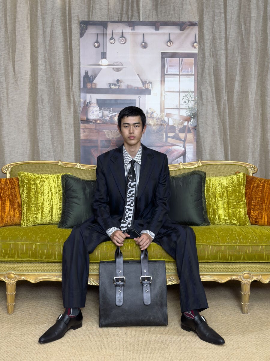 Man seated on a velvet sofa wearing a dark suit and branded tie, holding a black leather tote bag with silver buckles.