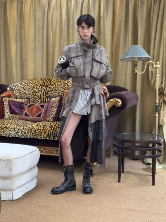Model wearing a layered plaid utility coat and lace-up combat boots standing by a leopard-print sofa with gold curtains.