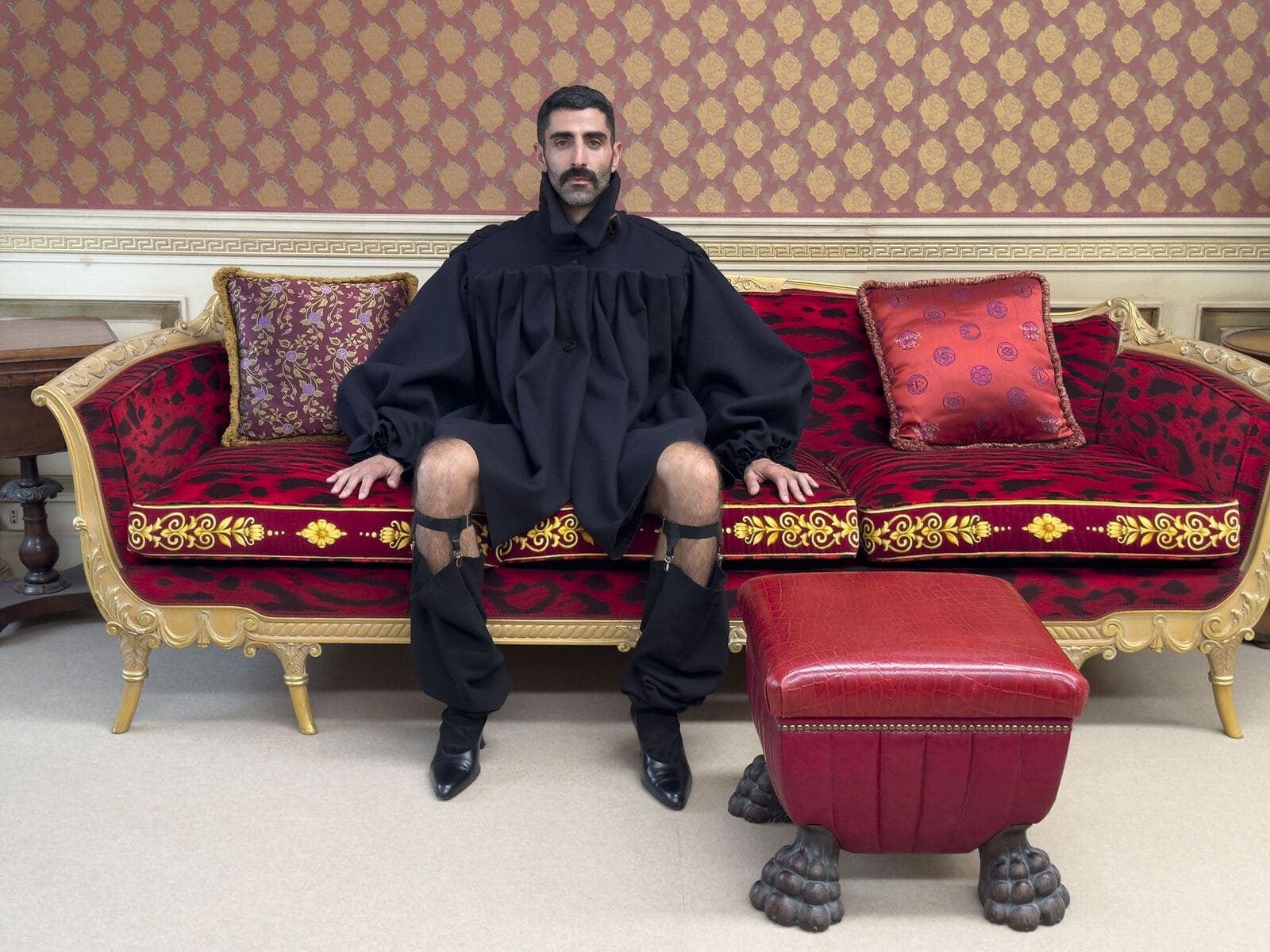 A man with a mustache seated on an ornate red and gold sofa wearing an oversized black garment and gartered footwear.
