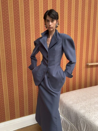 Model posing in a structured slate blue skirt suit with exaggerated shoulders in front of orange striped wallpaper.