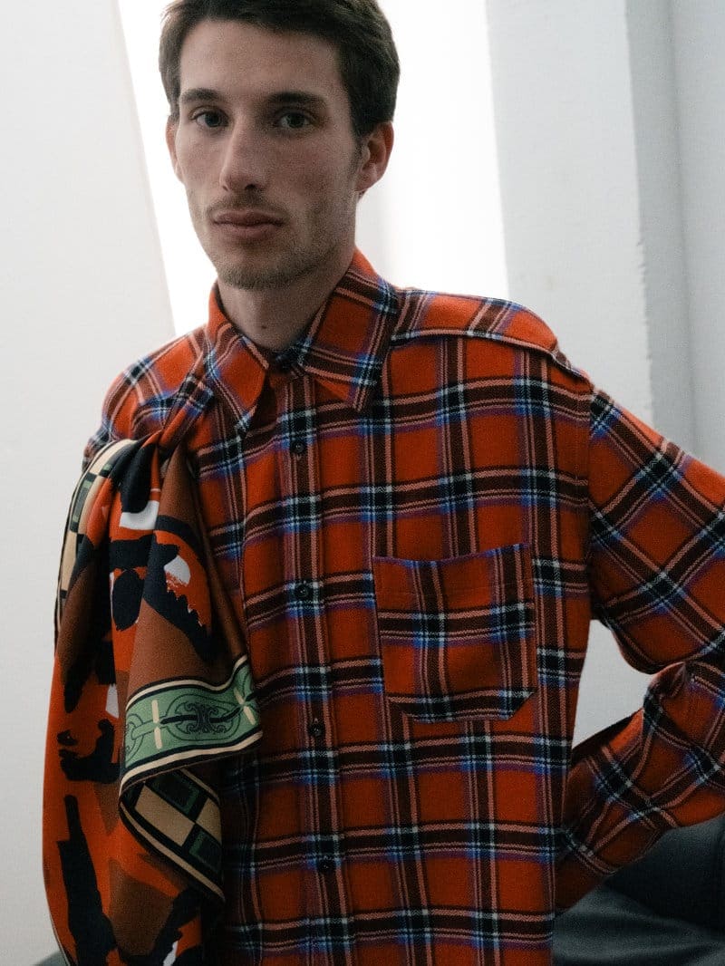 Man wearing an orange plaid shirt with a patterned scarf draped over his shoulder against a plain wall.