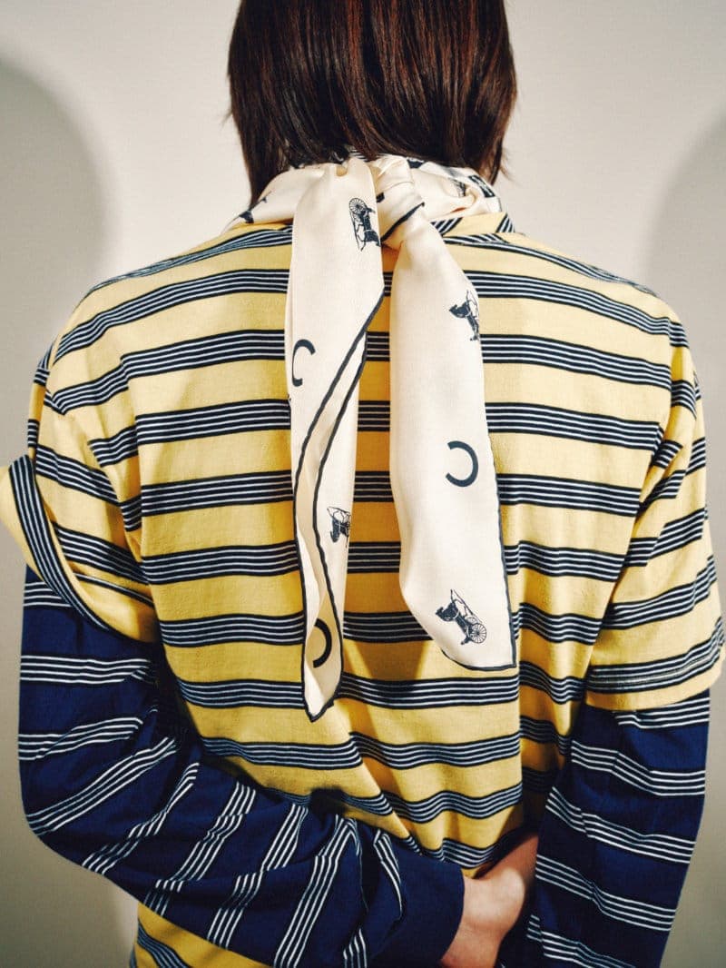 Person viewed from behind wearing layered yellow and navy striped shirts with a printed cream silk scarf knotted at the neck.