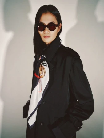 Model Haojie Qi wears dark round sunglasses, a black trench coat, and a graphic silk scarf against a pale studio wall.