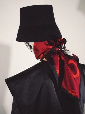 Fashion model viewed from behind wearing a tall black structured hat and a printed red silk scarf against a white background.