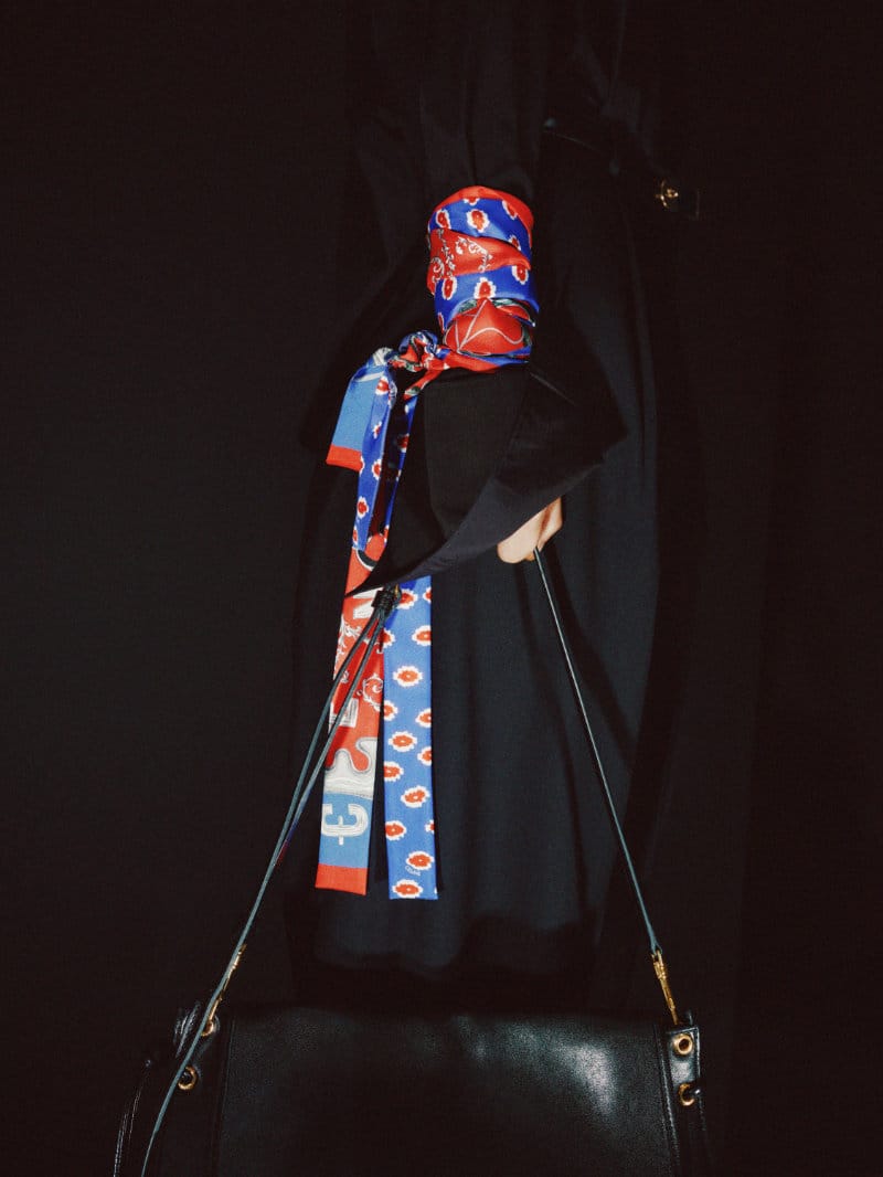 A wrist wrapped in a red and blue patterned silk scarf holding a black leather shoulder bag against a solid black background.