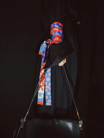A wrist wrapped in a red and blue patterned silk scarf holding a black leather shoulder bag against a solid black background.