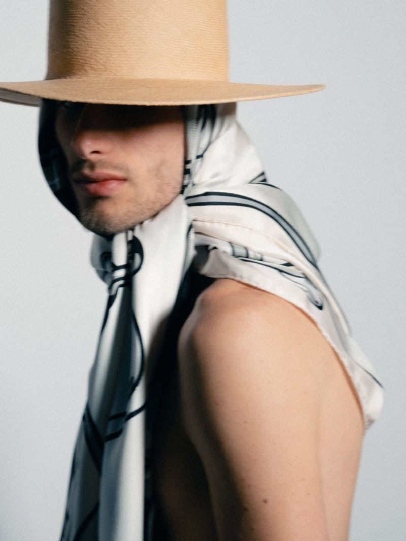 Man wearing a wide-brim tan hat and a draped printed silk scarf over bare shoulders against a white studio background.