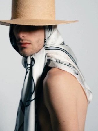 Man wearing a wide-brim tan hat and a draped printed silk scarf over bare shoulders against a white studio background.