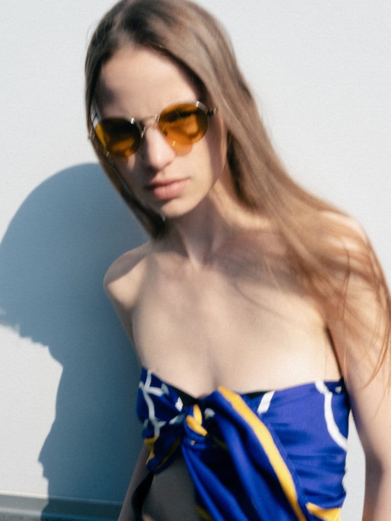 Model wearing yellow tinted sunglasses and a blue patterned top against a white wall with a sharp shadow cast