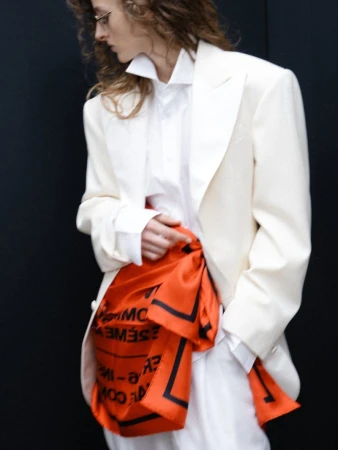 Model wearing an oversized cream tailored blazer, white shirt, and a bright orange printed scarf tied at the waist on a black background.